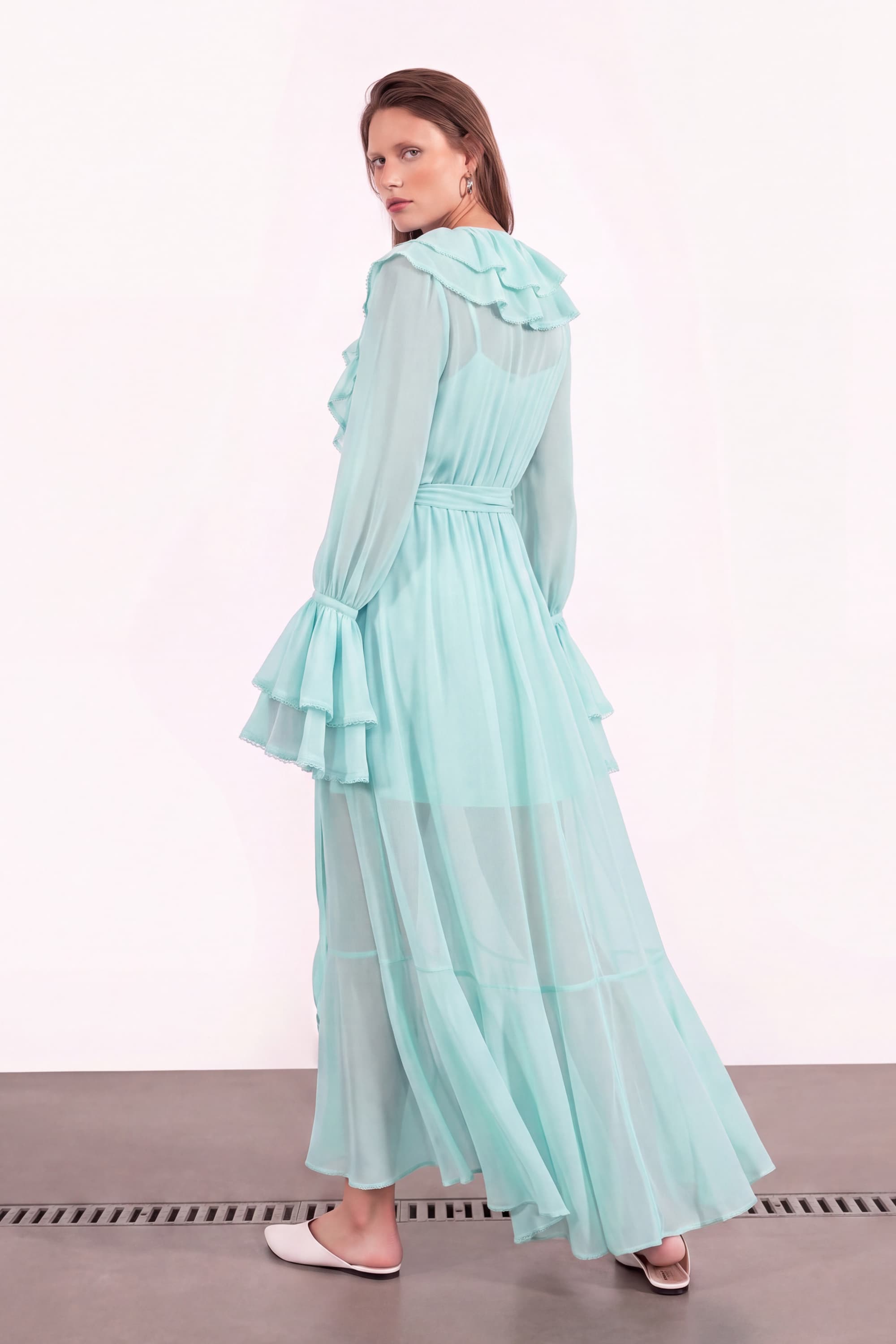 A woman modeling a long, tiered, ruffled maxi dress in a light mint green color, photographed from the back against a plain white studio background.