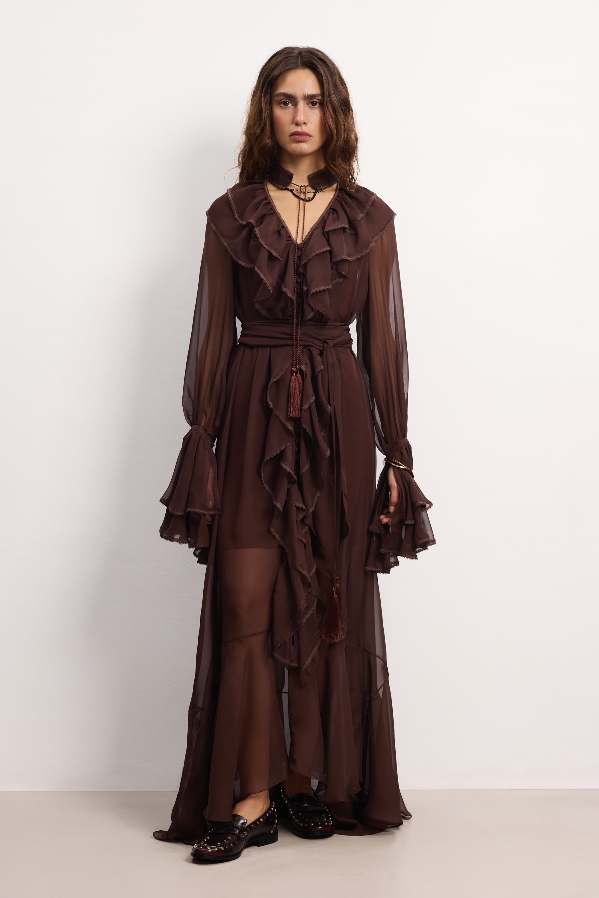 Model wearing a floor-length, sheer dark brown dress with cascading ruffles along the front, tiered sleeves, and a tassel choker