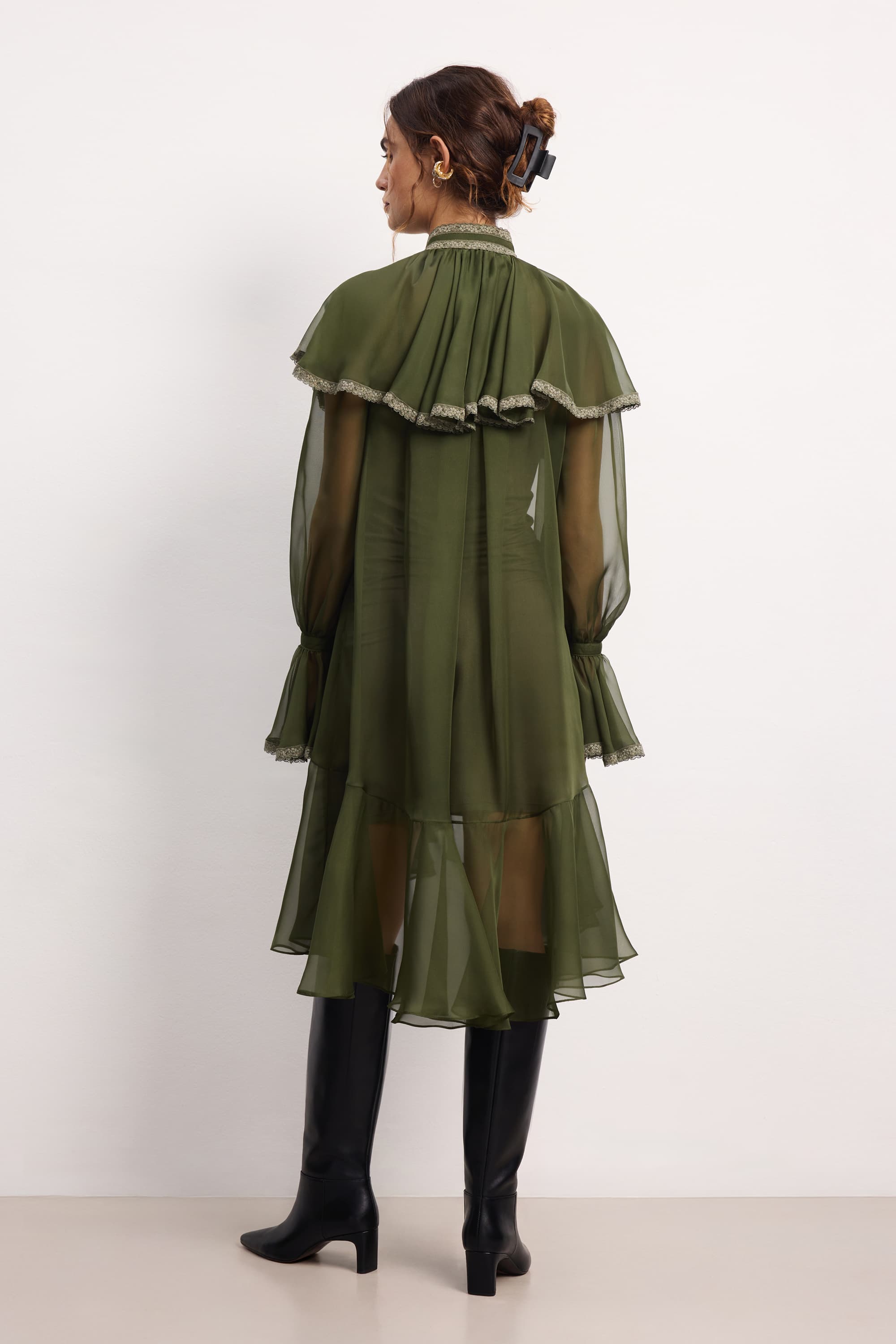 Woman wearing a olive ruffled Ocean Of Tenderness midi dress with black knee-high boots against a white background, back view